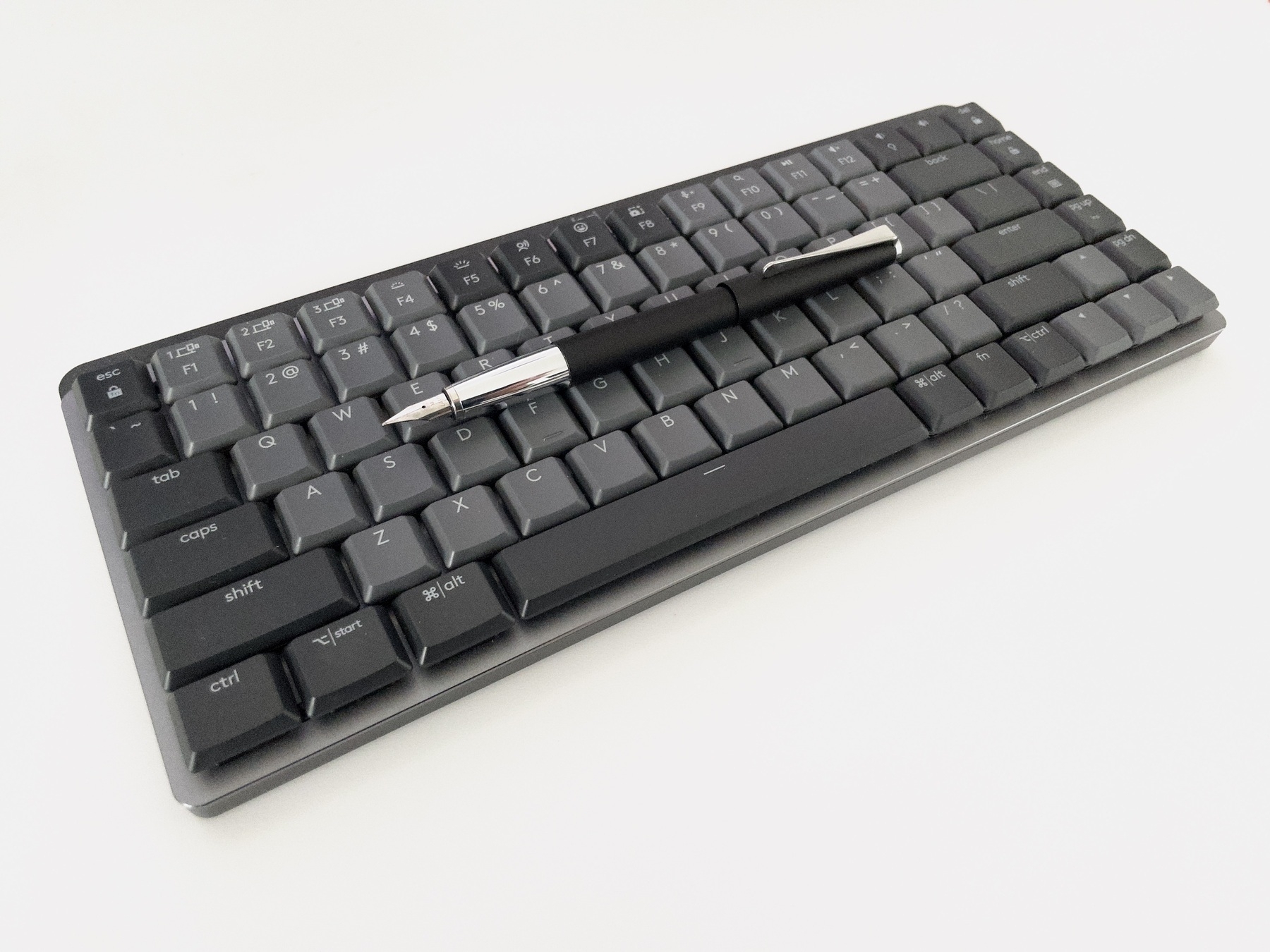 A Lamy Studio fountain pen in black with chrome accents, cap posted, sits atop a Logitech MX Mechanical Mini keyboard in dark grey. The background is a white desktop, giving the image a monochromatic feel without actually being black-and-white.