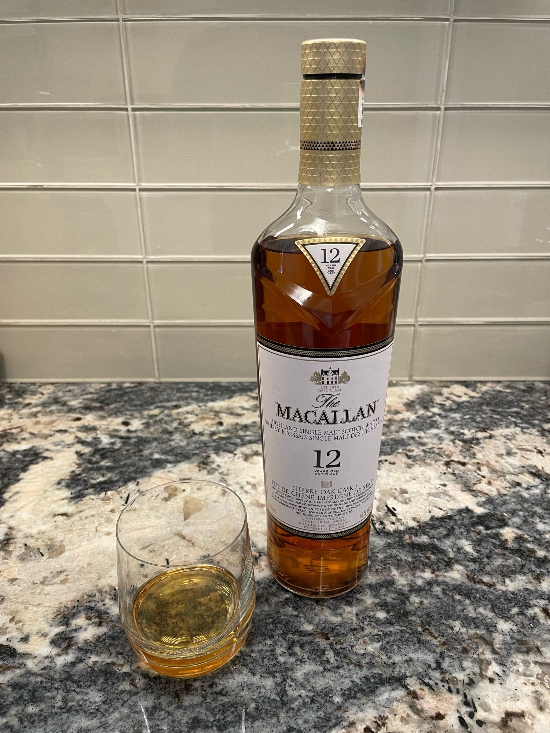 A bottle of Macallan sherry oak cask 12-year-old scotch next to a glass with a dram of it, both on a granite countertop.